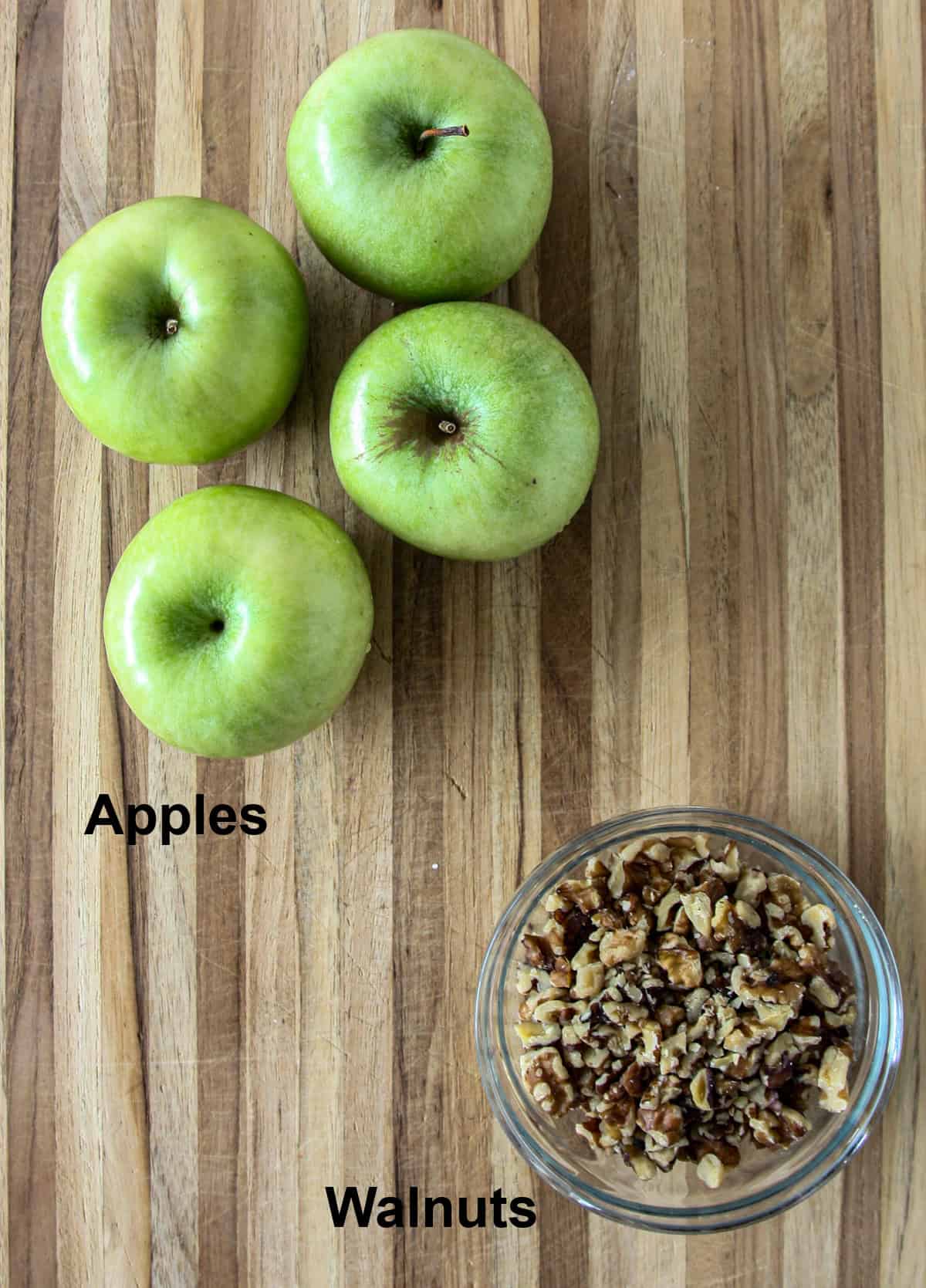 Four green apples and a dish of walnuts on a wooden board.