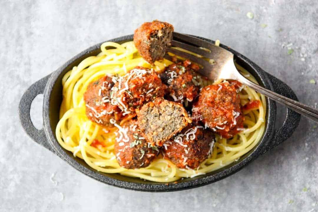 Top shot of fork holding half of meatless mushroom meatball above black bowl of spaghetti 