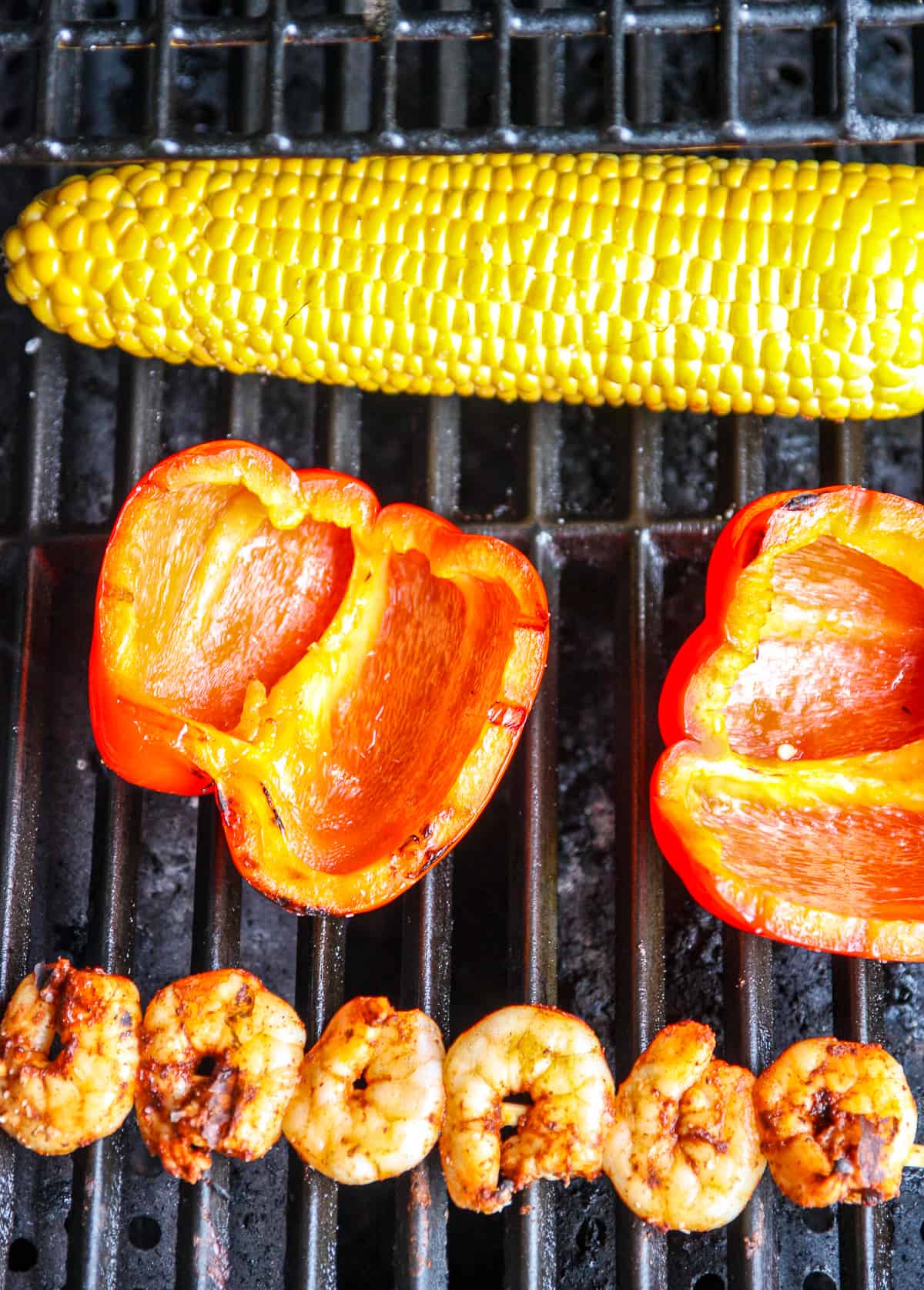 A cob of corn, a red bell pepper, and a skewer of shrimp on a grill. 