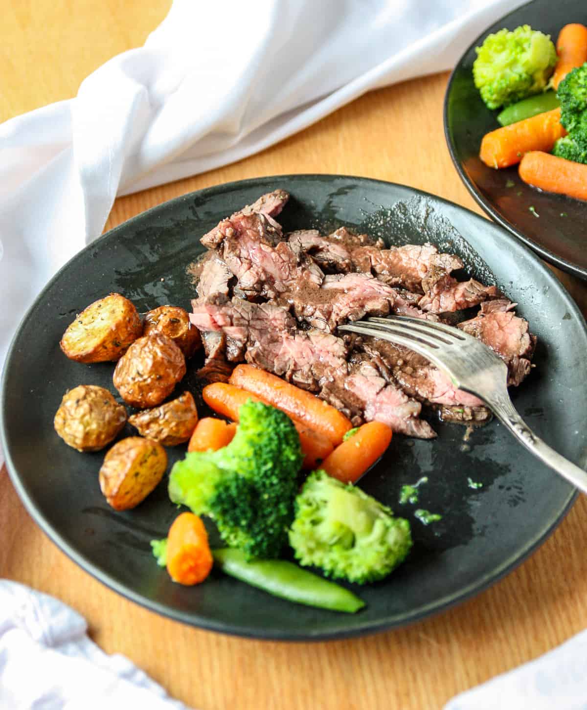 Sliced flank steak and roasted vegetables on a black plate with a fork.