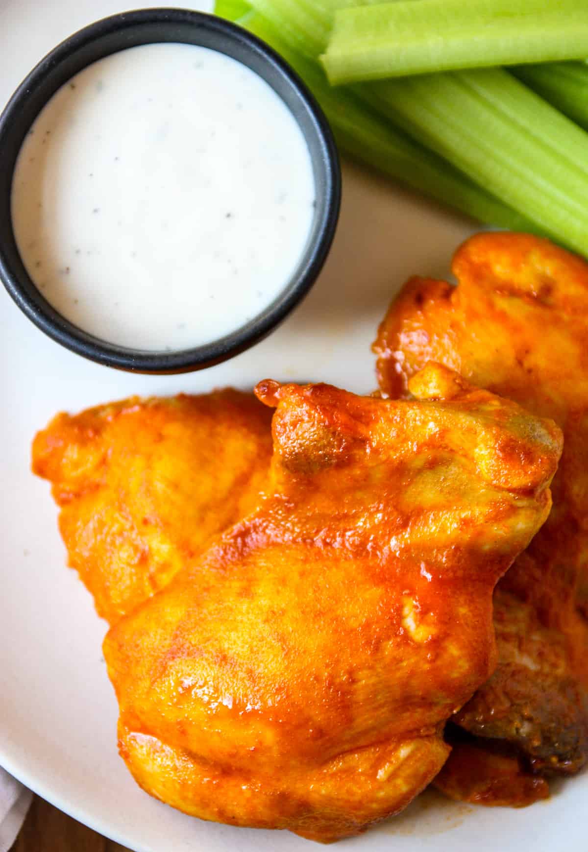 A white plate with buffalo chicken thighs, celery sticks and a small bowl of ranch dressing.