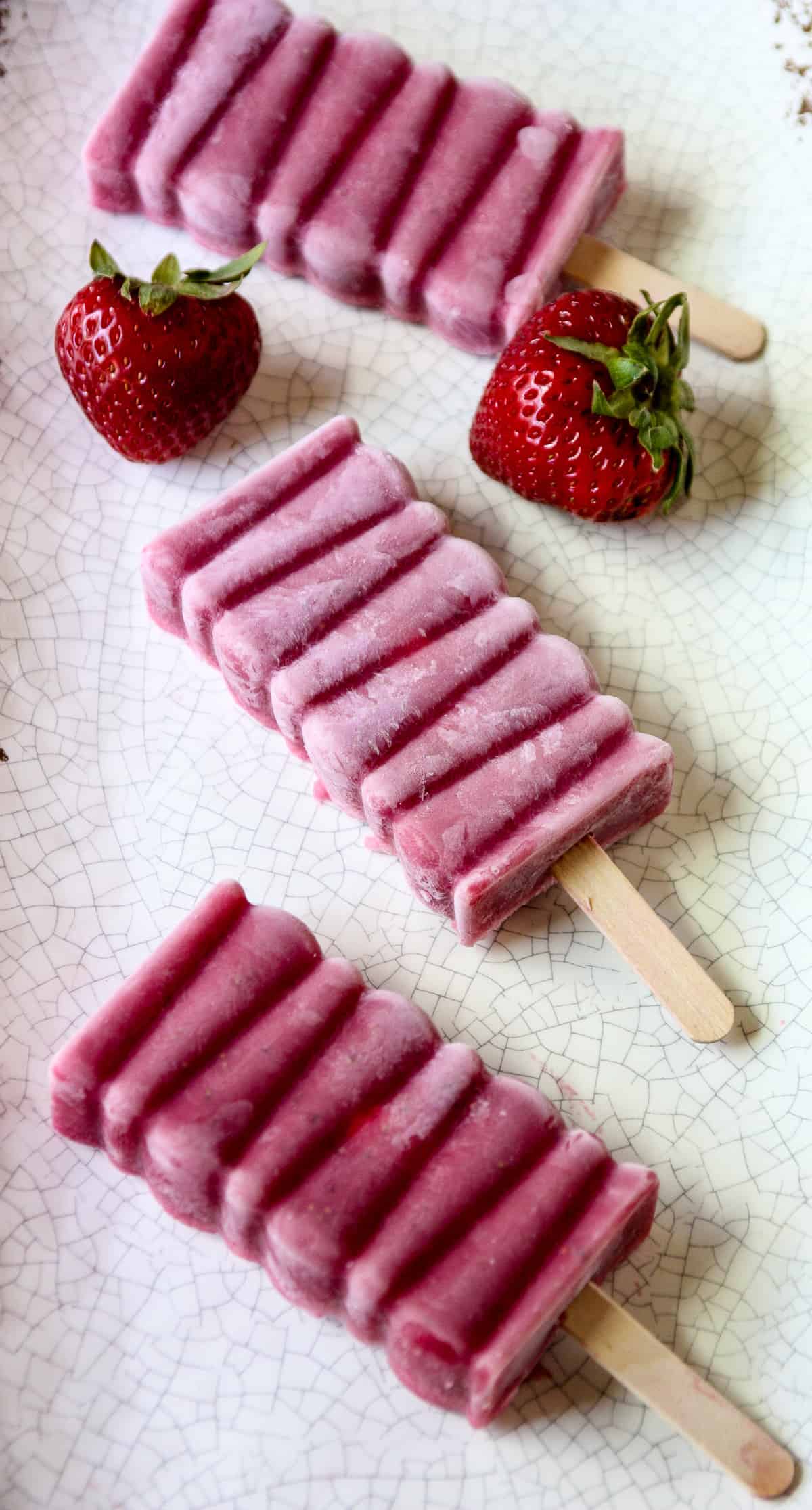 Two strawberry popsicles on a white speckled plate with two strawberries.
