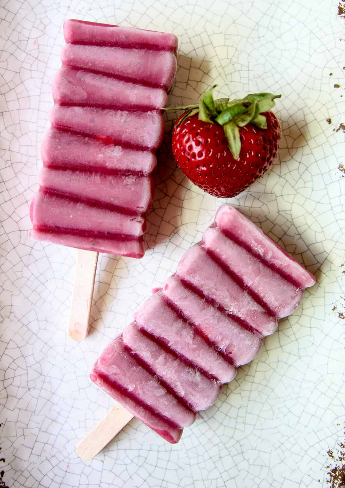 Homemade sugar free strawberry popsicles and a ripe strawberry on a speckled plate.