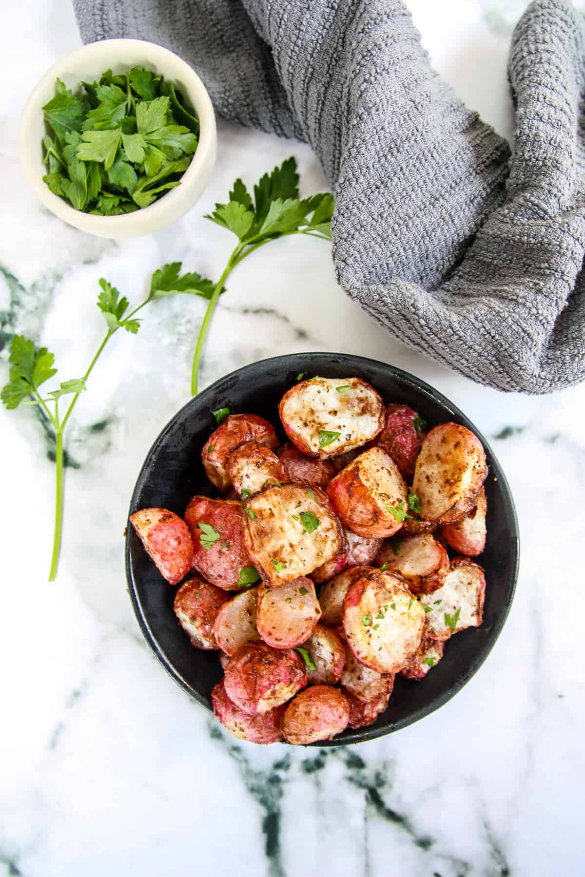 A black bowl filled with air fryer radishes next to a small white dish of parsley and a gray kitchen towel.