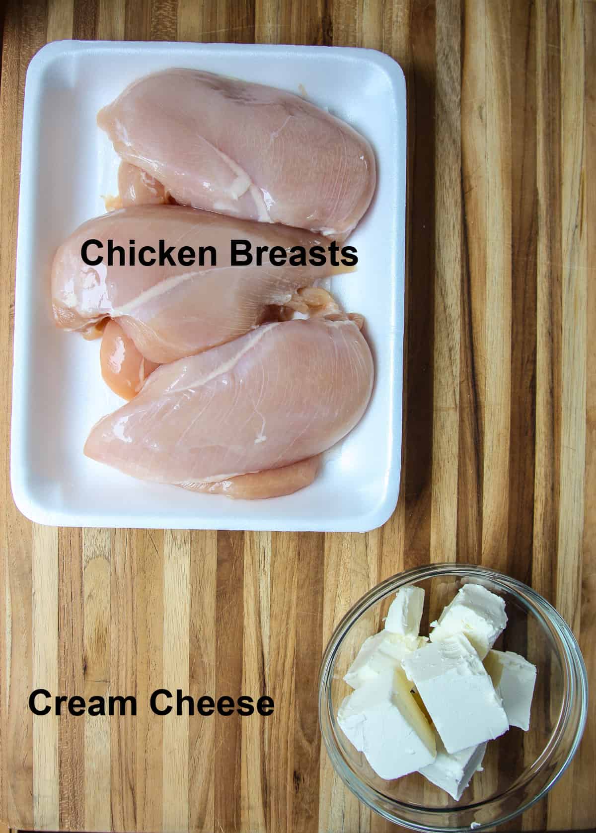 Uncooked chicken breasts and cubed cream cheese on a wooden board.