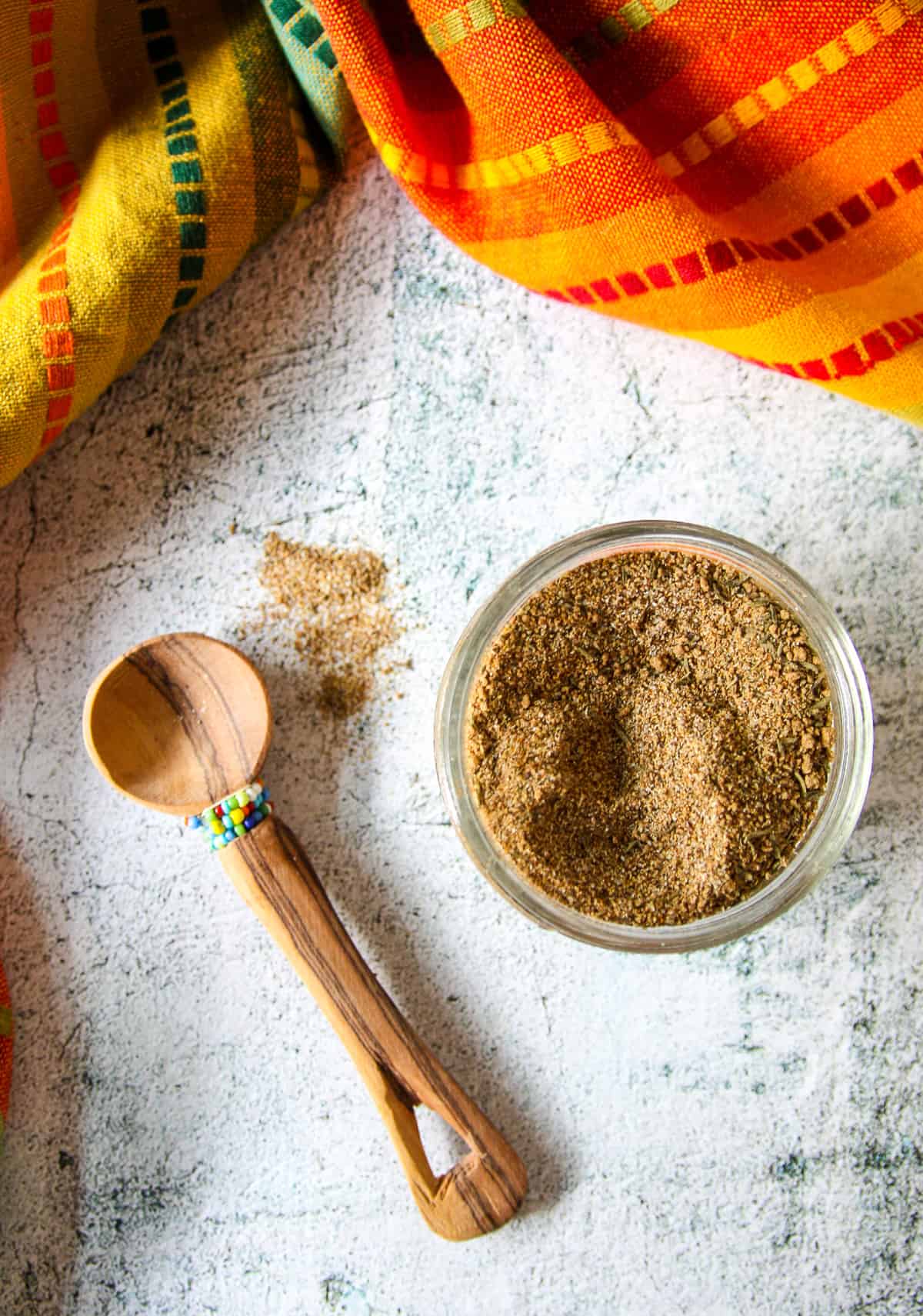 A small wooden spoon next to a glass jar full of Jamaican jerk seasoning blend.