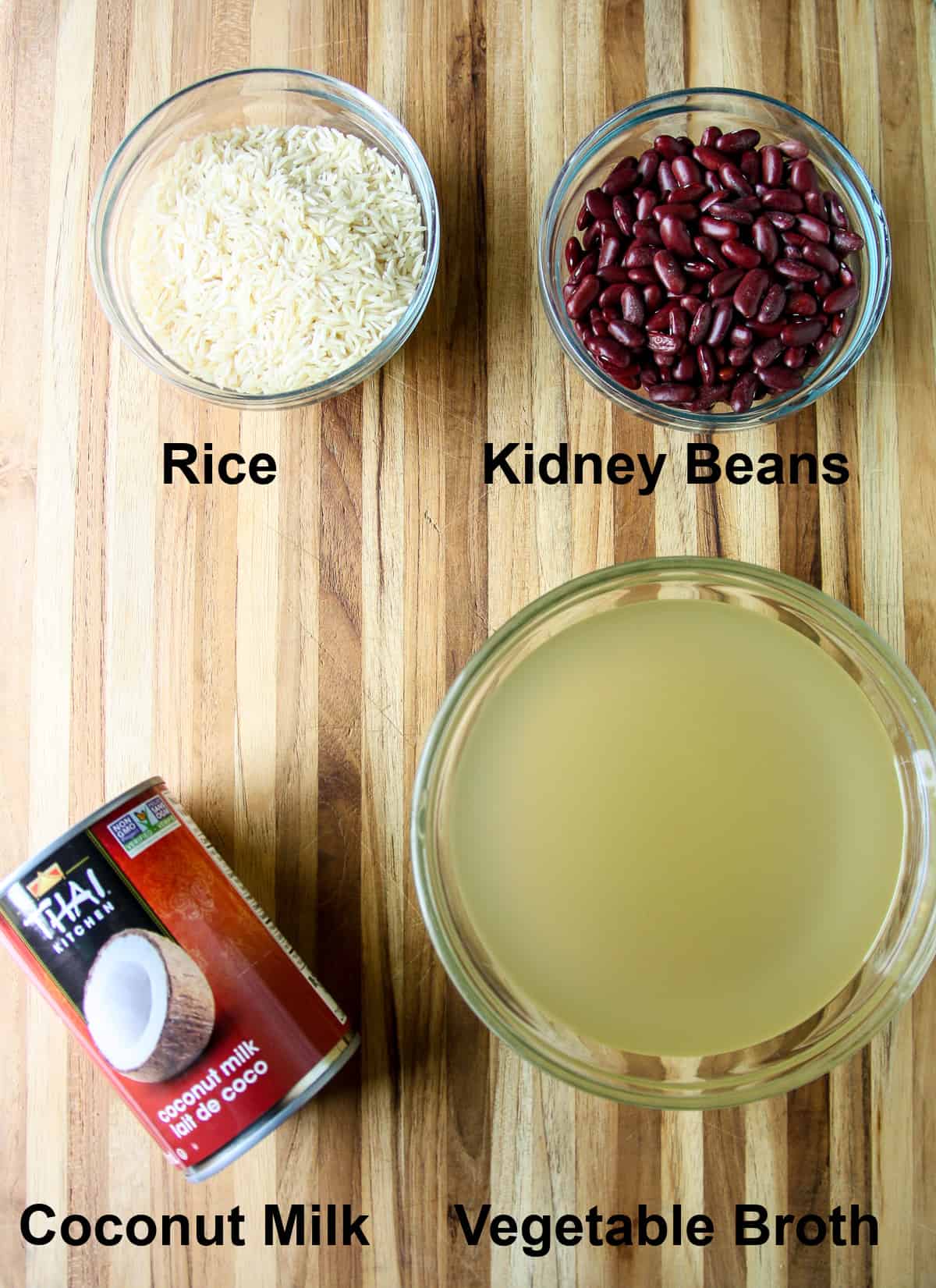 Uncooked rice, kidney beans, coconut milk, and vegetable broth in glass bowls on a wooden board.