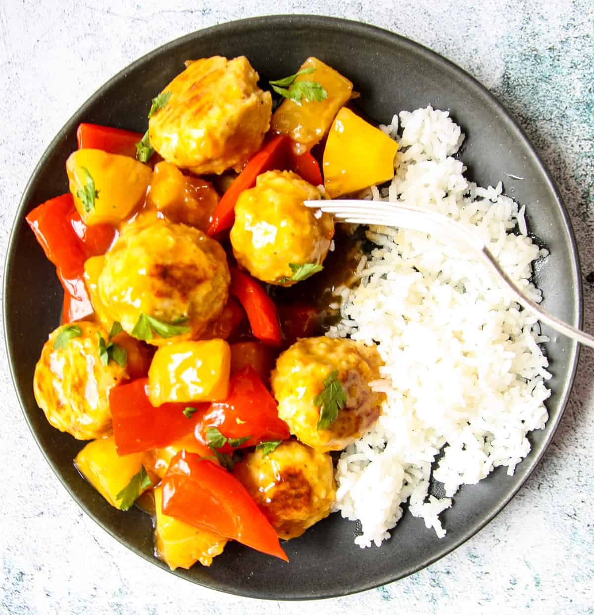 A silver fork dipping into a plate full of sweet and sour meatballs and rice.