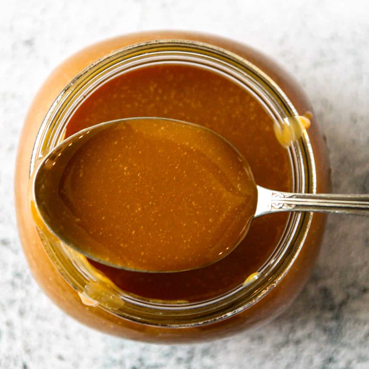 A spoon full of caramel candy sauce resting on the rim of a jar.